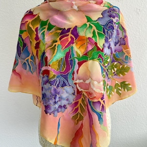 May include: A square silk scarf featuring a hand-painted floral design with vibrant shades of pink, purple, green, orange, and blue. The pattern displays large blossoms and foliage with defined outlines, set against a soft peach and cream background.