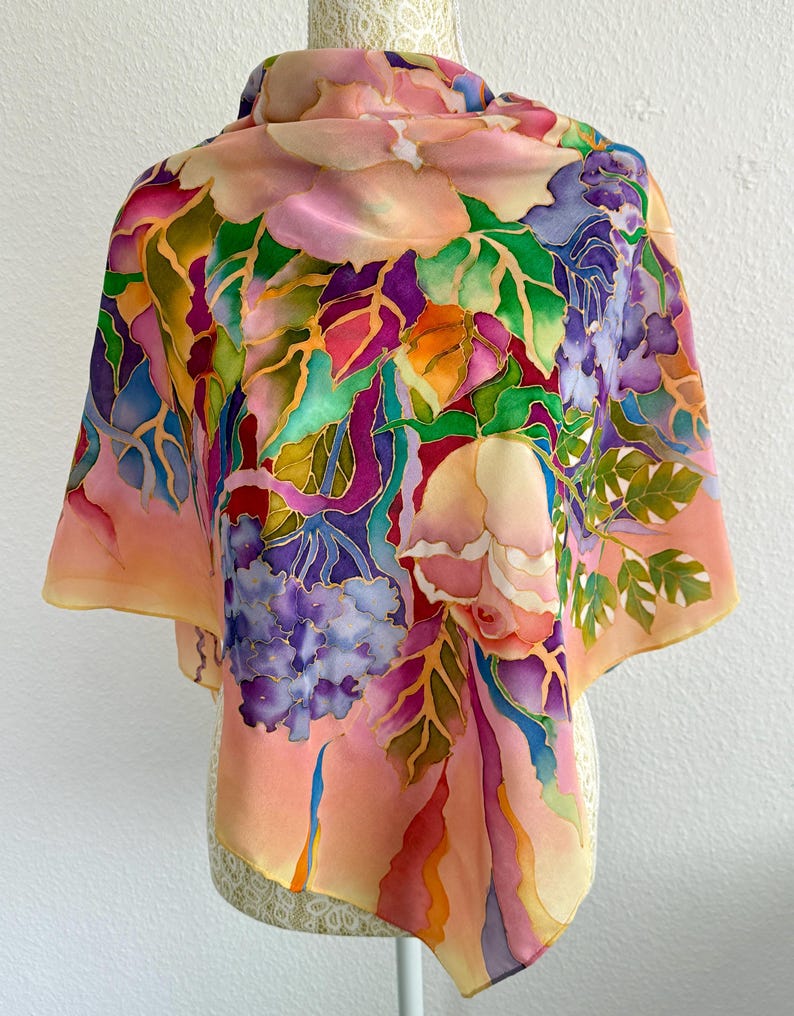 May include: A square silk scarf featuring a hand-painted floral design with vibrant shades of pink, purple, green, orange, and blue. The pattern displays large blossoms and foliage with defined outlines, set against a soft peach and cream background.
