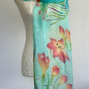 Pode incluir: Um len&ccedil;o leve e semitransparente num tom turquesa vibrante, com um design floral pintado &agrave; m&atilde;o com flores cor de laranja, rosa e amarelas. O len&ccedil;o apresenta caules e folhas verdes delicados, com um caimento suave e uma textura lisa.