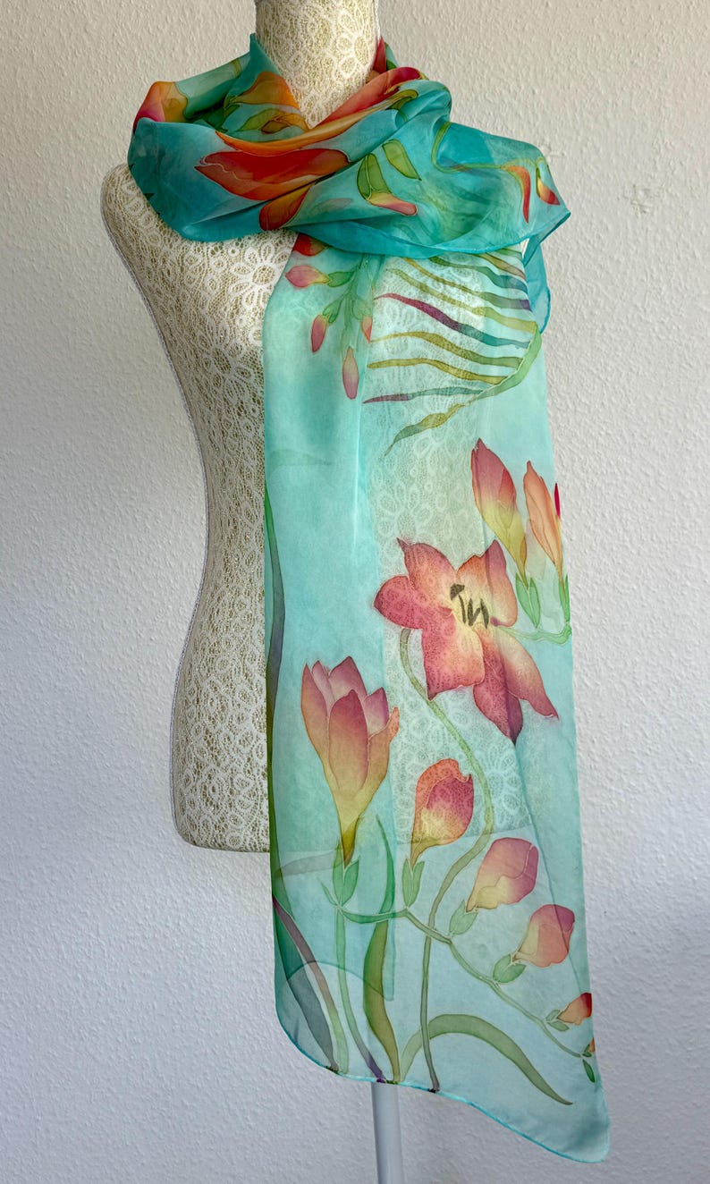 Pode incluir: Um len&ccedil;o leve e semitransparente num tom turquesa vibrante, com um design floral pintado &agrave; m&atilde;o com flores cor de laranja, rosa e amarelas. O len&ccedil;o apresenta caules e folhas verdes delicados, com um caimento suave e uma textura lisa.