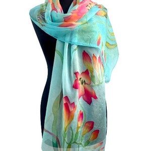 Pode incluir: Um len&ccedil;o leve e semitransparente num tom azul turquesa vibrante, com um padr&atilde;o floral pintado &agrave; m&atilde;o que apresenta flores em tons de rosa brilhante, coral e laranja, acentuadas por caules e folhas verdes finas ao longo do tecido.