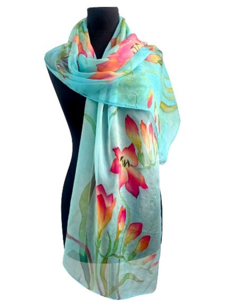 Pode incluir: Um len&ccedil;o leve e semitransparente num tom azul turquesa vibrante, com um padr&atilde;o floral pintado &agrave; m&atilde;o que apresenta flores em tons de rosa brilhante, coral e laranja, acentuadas por caules e folhas verdes finas ao longo do tecido.