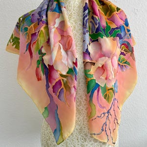 May include: A square silk scarf featuring a hand-painted floral pattern in shades of peach, pink, purple, blue, and green. The design includes large blossoms and leaves with defined outlines, set against a soft peach background with blended colors.