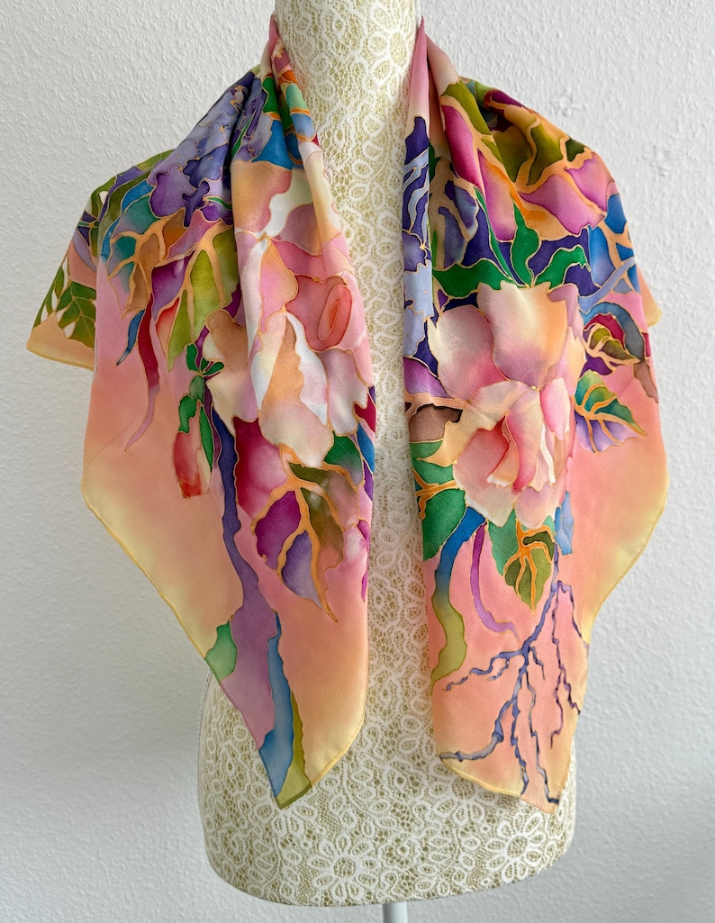 May include: A square silk scarf featuring a hand-painted floral pattern in shades of peach, pink, purple, blue, and green. The design includes large blossoms and leaves with defined outlines, set against a soft peach background with blended colors.
