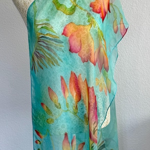 Pode incluir: Um len&ccedil;o leve e semitransparente com um fundo turquesa vibrante e um padr&atilde;o branco subtil e repetitivo semelhante a renda. O len&ccedil;o &eacute; decorado com motivos florais pintados &agrave; m&atilde;o em tons de laranja, rosa e verde, representando flores.