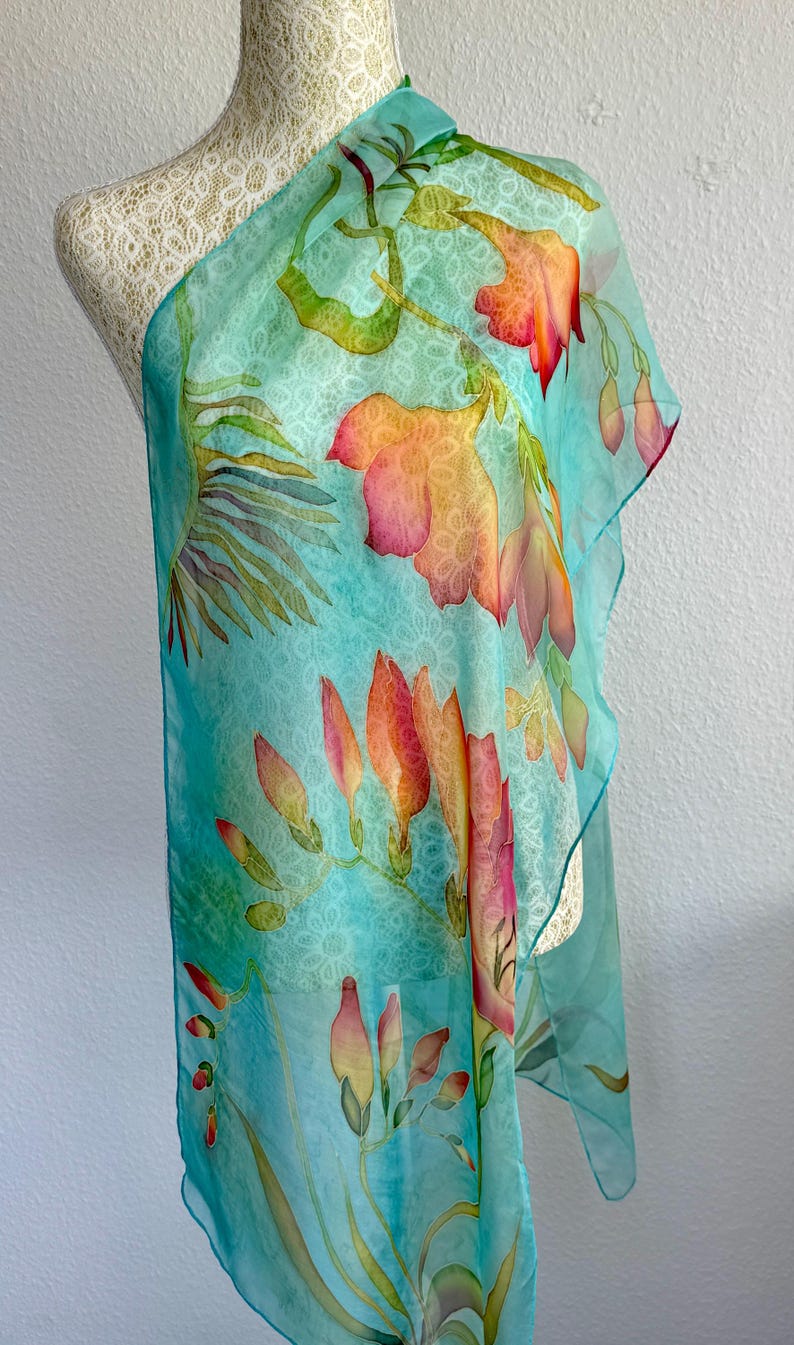 Pode incluir: Um len&ccedil;o leve e semitransparente com um fundo turquesa vibrante e um padr&atilde;o branco subtil e repetitivo semelhante a renda. O len&ccedil;o &eacute; decorado com motivos florais pintados &agrave; m&atilde;o em tons de laranja, rosa e verde, representando flores.