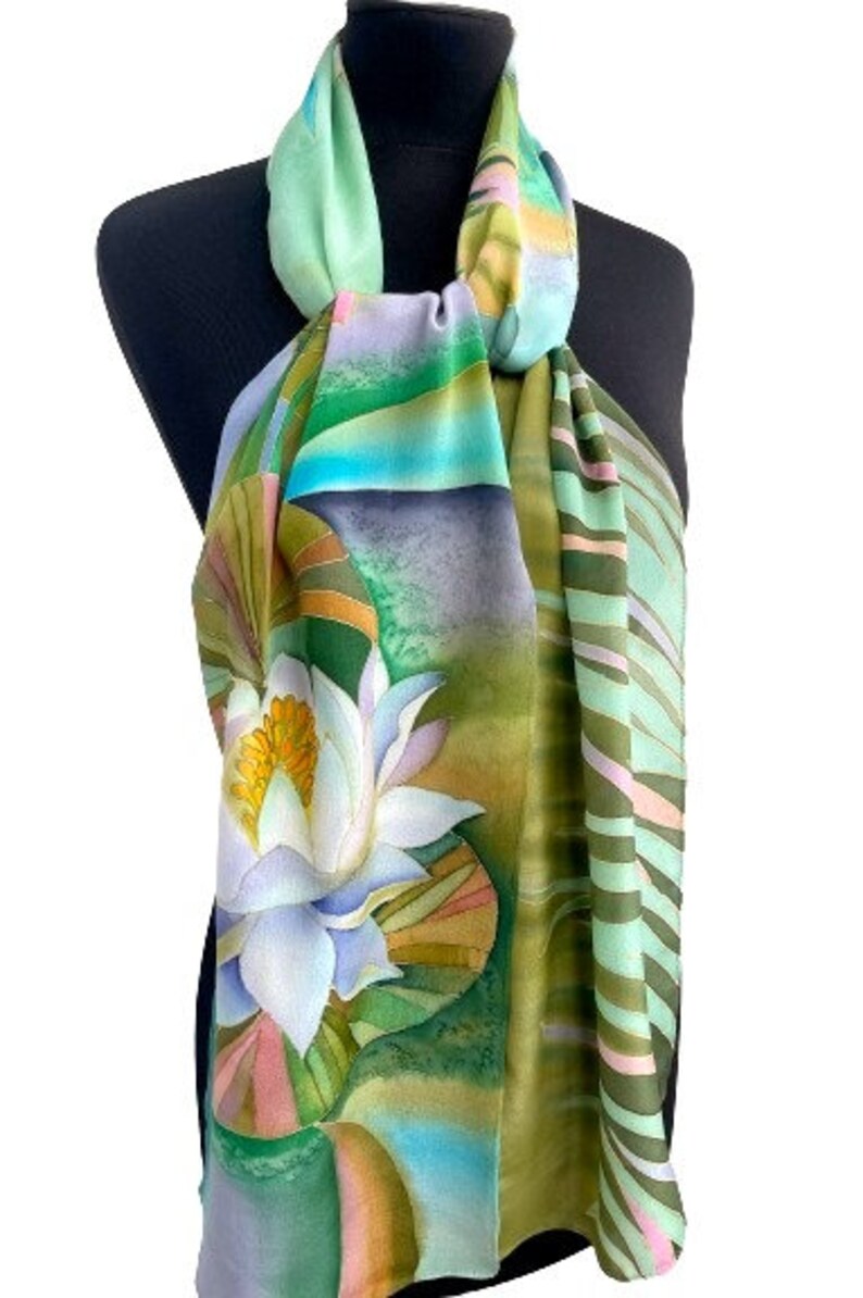 Puede incluir: Una bufanda de seda verde, azul y blanca con un dise&ntilde;o pintado a mano de un nen&uacute;far. La bufanda tiene un patr&oacute;n de rayas en un lado y un patr&oacute;n floral en el otro.