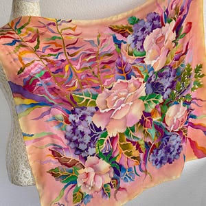 May include: A square silk scarf featuring a hand-painted floral design on a peach background. The pattern includes large pink roses, clusters of purple hydrangeas, and green fern-like foliage with vibrant accents of blue, yellow, and magenta.