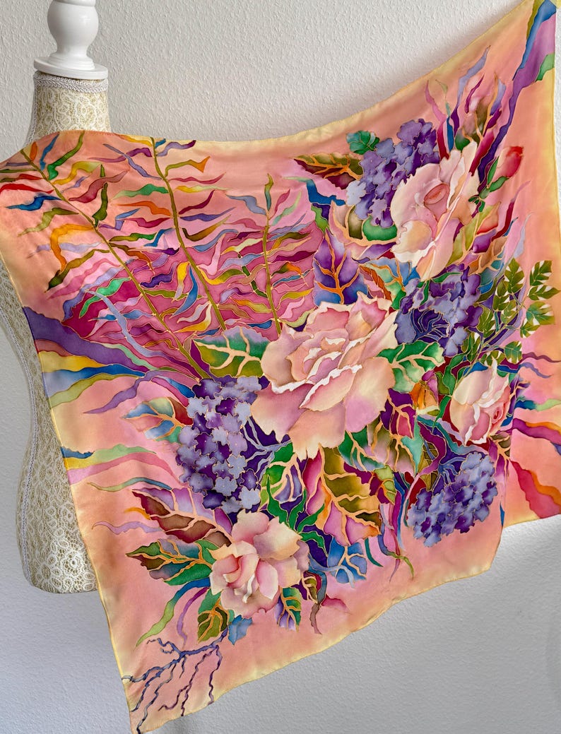 May include: A square silk scarf featuring a hand-painted floral design on a peach background. The pattern includes large pink roses, clusters of purple hydrangeas, and green fern-like foliage with vibrant accents of blue, yellow, and magenta.