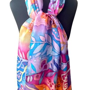 May include: A colorful silk scarf with a floral and abstract design in shades of pink, blue, orange, and yellow. The scarf is tied around a mannequin's neck.