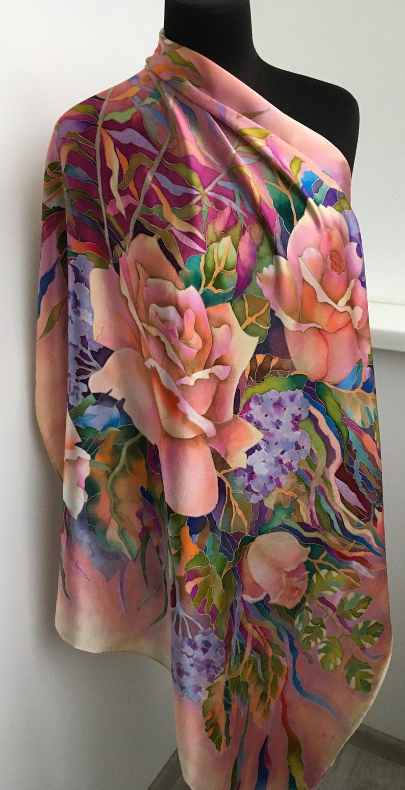 May include: A silk scarf featuring a hand-painted floral pattern with large peach-colored roses, clusters of small purple flowers, and green foliage. The background transitions between shades of soft pink, deep magenta, and violet, creating a vibrant, multi-colored botanical design on the fabric.