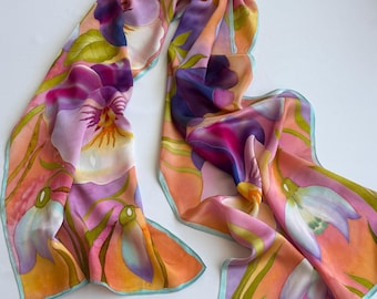 Puede incluir: Una bufanda larga y rectangular de seda con un estampado floral pintado a mano que presenta grandes flores moradas, rosas y blancas sobre un fondo degradado de color melocotón y naranja. Los bordes tienen un dobladillo azul claro y tallos verdes.