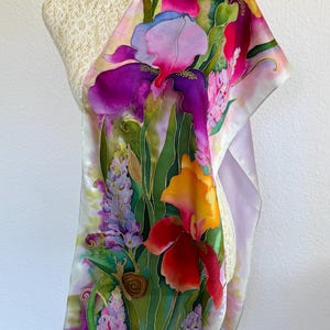 K&ouml;nnte beinhalten: Ein rechteckiger Seidenschal mit einem lebendigen, handgemalten Blumendesign mit gro&szlig;en violetten, orangefarbenen und roten Schwertlilien, Lavendelst&auml;ngeln und gr&uuml;nem Blattwerk. Eine kleine braune Schnecke ist unten links zwischen den Blumen zu sehen.