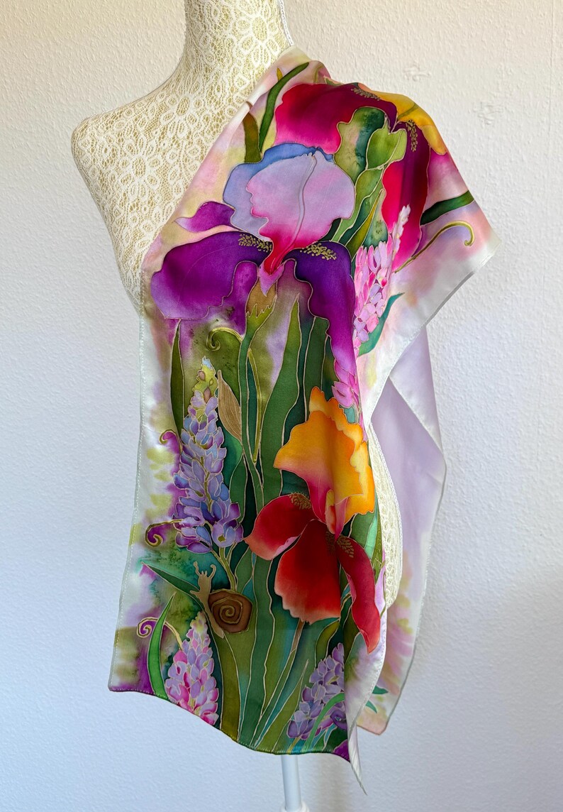 K&ouml;nnte beinhalten: Ein rechteckiger Seidenschal mit einem lebendigen, handgemalten Blumendesign mit gro&szlig;en violetten, orangefarbenen und roten Schwertlilien, Lavendelst&auml;ngeln und gr&uuml;nem Blattwerk. Eine kleine braune Schnecke ist unten links zwischen den Blumen zu sehen.