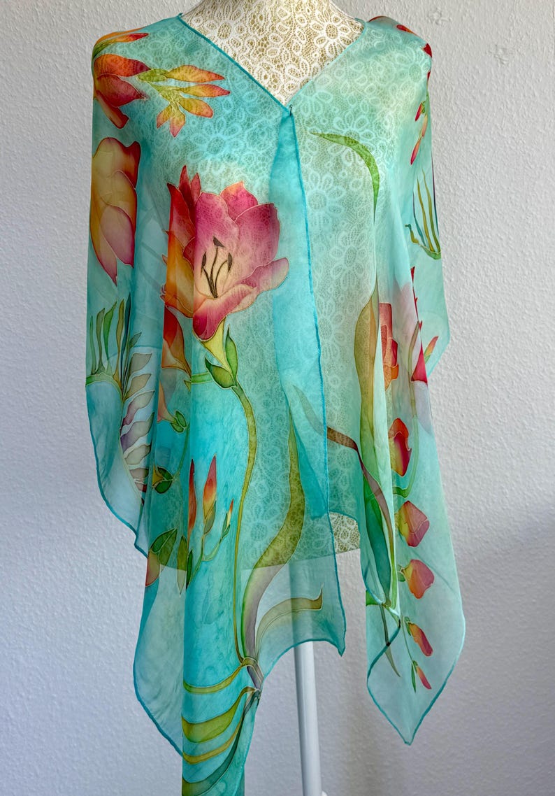 Pode incluir: Um len&ccedil;o leve e semitransparente num tom turquesa vibrante, com um delicado padr&atilde;o branco semelhante a renda. O tecido &eacute; adornado com motivos florais pintados &agrave; m&atilde;o em tons de rosa, laranja e amarelo, com caules verdes.
