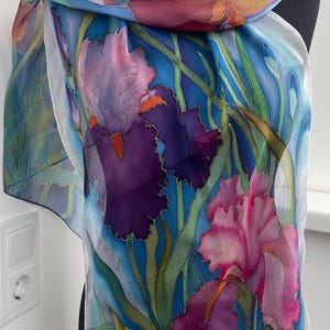 May include: A lightweight silk scarf featuring a hand-painted floral design of purple, pink, and orange iris flowers with green stems against a vibrant blue background. The scarf has a smooth, flowing texture and includes a signature on the lower left.