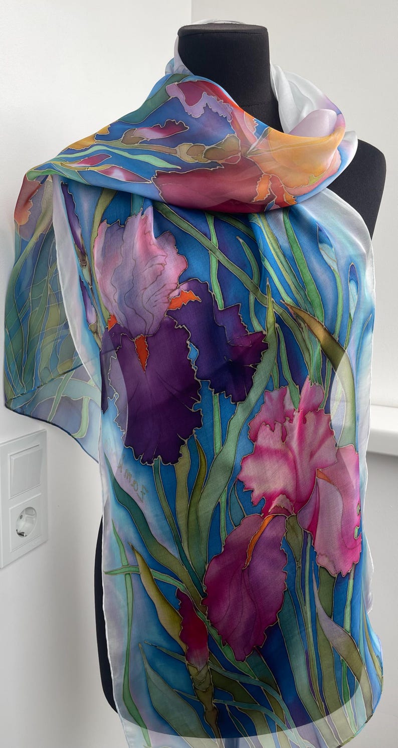 May include: A lightweight silk scarf featuring a hand-painted floral design of purple, pink, and orange iris flowers with green stems against a vibrant blue background. The scarf has a smooth, flowing texture and includes a signature on the lower left.