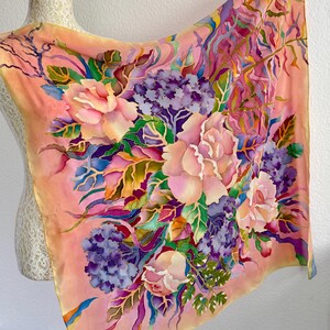May include: A square silk scarf featuring a hand-painted floral design on a peach background. The pattern includes large pink roses, clusters of purple hydrangeas, and green foliage with blue and yellow accents, creating a vibrant botanical display.