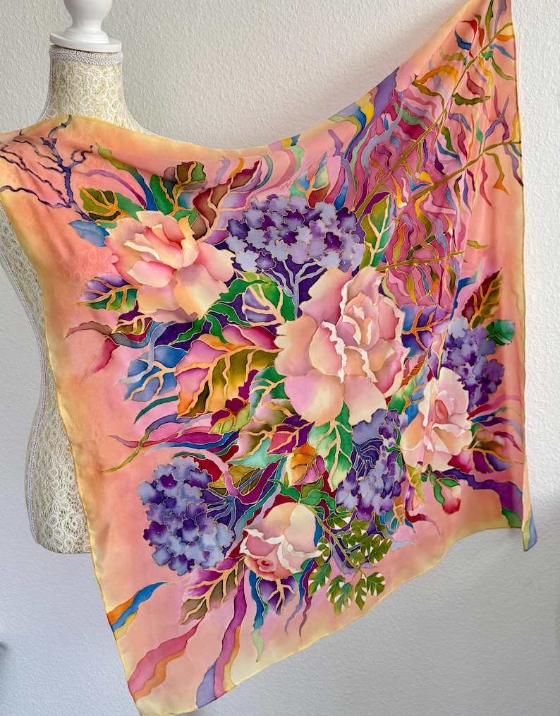 May include: A square silk scarf featuring a hand-painted floral design on a peach background. The pattern includes large pink roses, clusters of purple hydrangeas, and green foliage with blue and yellow accents, creating a vibrant botanical display.