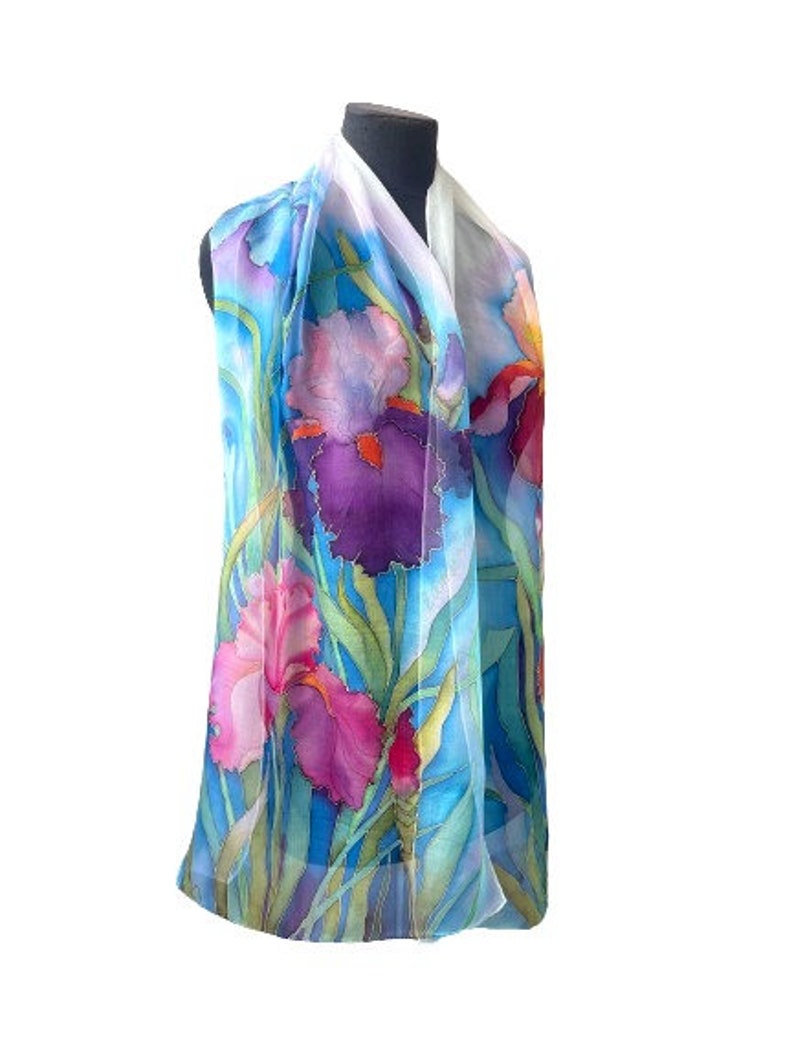 May include: A lightweight scarf featuring a hand-painted floral design of purple, pink, and deep red iris flowers set against a vibrant blue background with long green stems and leaves, displayed on a black mannequin torso for clear visibility.