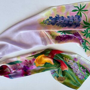 K&ouml;nnte beinhalten: Ein langer, rechteckiger Seidenschal mit handgemaltem Blumenmuster. Der Stoff zeigt eine zarte lavendelfarbene Grundfarbe mit lebendigen Illustrationen von blauen Lupinen, gelben Tulpen und gr&uuml;nem Blattwerk, akzentuiert durch ein kleines braunes Schneckendetail.