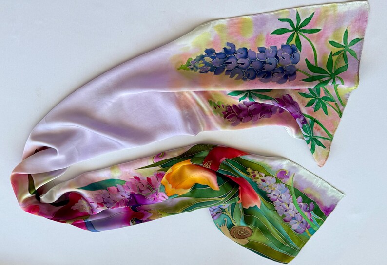 K&ouml;nnte beinhalten: Ein langer, rechteckiger Seidenschal mit handgemaltem Blumenmuster. Der Stoff zeigt eine zarte lavendelfarbene Grundfarbe mit lebendigen Illustrationen von blauen Lupinen, gelben Tulpen und gr&uuml;nem Blattwerk, akzentuiert durch ein kleines braunes Schneckendetail.