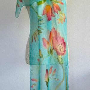 Pode incluir: Um len&ccedil;o retangular num tom azul turquesa claro com um padr&atilde;o floral pintado &agrave; m&atilde;o. O design apresenta grandes e vibrantes flores cor de laranja e rosa com caules e folhas verdes, distribu&iacute;das pelo tecido leve e transparente.