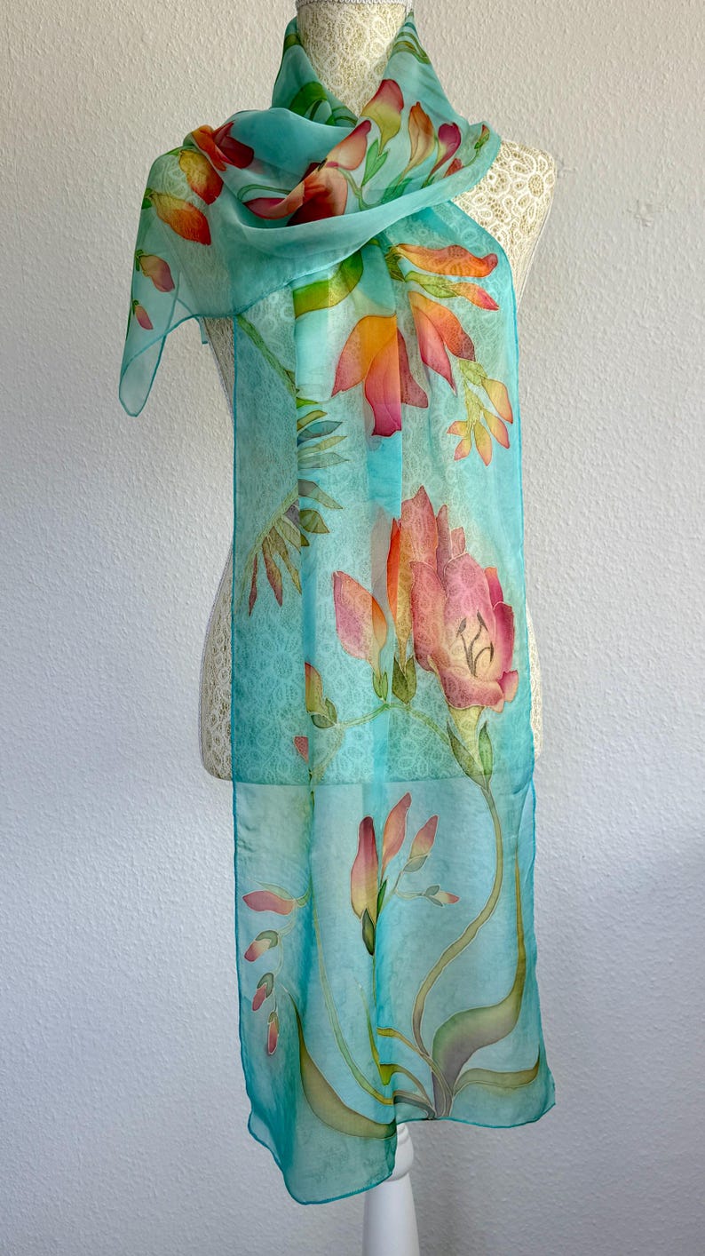Pode incluir: Um len&ccedil;o retangular num tom azul turquesa claro com um padr&atilde;o floral pintado &agrave; m&atilde;o. O design apresenta grandes e vibrantes flores cor de laranja e rosa com caules e folhas verdes, distribu&iacute;das pelo tecido leve e transparente.
