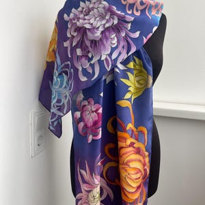 May include: A silk scarf with a blue background and a colorful floral pattern. The flowers are painted in shades of purple, yellow, orange, and pink. The scarf is draped over a mannequin.