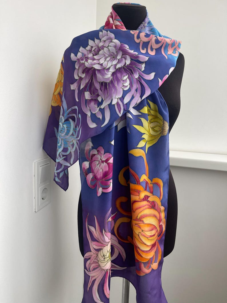 May include: A silk scarf with a blue background and a colorful floral pattern. The flowers are painted in shades of purple, yellow, orange, and pink. The scarf is draped over a mannequin.