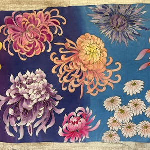 May include: A silk scarf with a vibrant floral pattern. The scarf features a variety of chrysanthemum flowers in shades of yellow, pink, purple, and blue, as well as smaller white flowers. The background of the scarf is a gradient of purple and blue.
