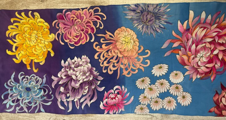 May include: A silk scarf with a vibrant floral pattern. The scarf features a variety of chrysanthemum flowers in shades of yellow, pink, purple, and blue, as well as smaller white flowers. The background of the scarf is a gradient of purple and blue.