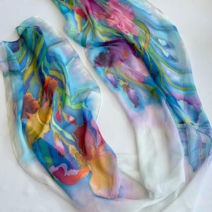 May include: Two lightweight silk scarves featuring hand-painted floral designs. The fabric displays vibrant iris flowers in shades of pink, purple, orange, and yellow, set against a background of blue and green foliage on a white base.