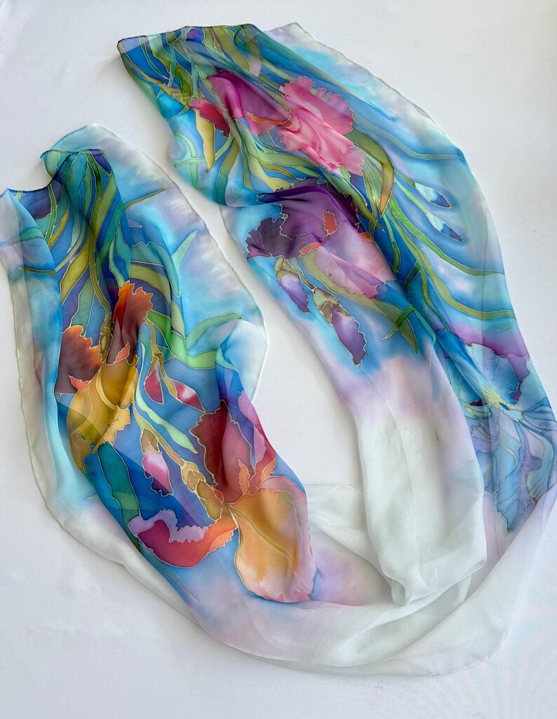 May include: Two lightweight silk scarves featuring hand-painted floral designs. The fabric displays vibrant iris flowers in shades of pink, purple, orange, and yellow, set against a background of blue and green foliage on a white base.