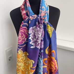 May include: A silk scarf with a vibrant floral pattern featuring chrysanthemums in shades of pink, purple, yellow, and orange on a blue and purple gradient background.