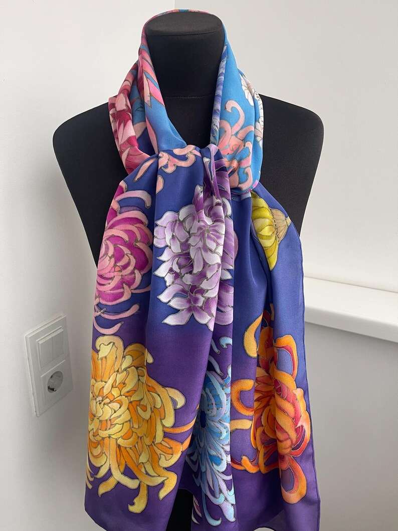 May include: A silk scarf with a vibrant floral pattern featuring chrysanthemums in shades of pink, purple, yellow, and orange on a blue and purple gradient background.