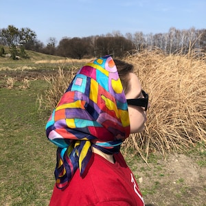 May include: A person wearing a colourful silk scarf with geometric patterns in shades of red, yellow, blue, purple, and green. The scarf is tied around the head and covers the hair. The person is wearing sunglasses and a red shirt.