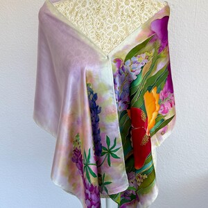 K&ouml;nnte beinhalten: Ein rechteckiger Seidenschal mit handgemaltem Blumenmuster, das leuchtend violette Lupinen, rote und orangefarbene hibiskusartige Bl&uuml;ten sowie gr&uuml;nes Blattwerk auf einem zart lavendelfarbenen und wei&szlig;en Hintergrund zeigt, drapiert &uuml;ber einer wei&szlig;en Spitzenpuppe.