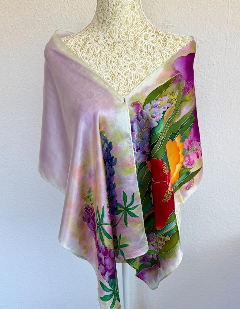 K&ouml;nnte beinhalten: Ein rechteckiger Seidenschal mit handgemaltem Blumenmuster, das leuchtend violette Lupinen, rote und orangefarbene hibiskusartige Bl&uuml;ten sowie gr&uuml;nes Blattwerk auf einem zart lavendelfarbenen und wei&szlig;en Hintergrund zeigt, drapiert &uuml;ber einer wei&szlig;en Spitzenpuppe.