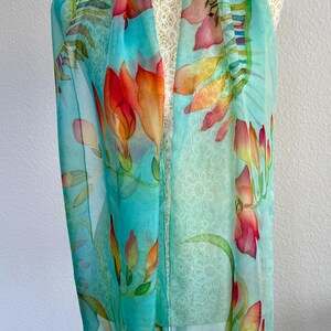 Pode incluir: Um len&ccedil;o leve e semitransparente com um fundo turquesa vibrante adornado com motivos florais pintados &agrave; m&atilde;o em tons de laranja, vermelho e amarelo. O design inclui caules e folhas verdes delicados, criando um padr&atilde;o bot&acirc;nico.