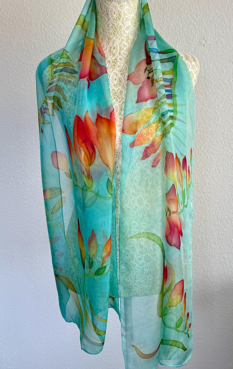 Pode incluir: Um len&ccedil;o leve e semitransparente com um fundo turquesa vibrante adornado com motivos florais pintados &agrave; m&atilde;o em tons de laranja, vermelho e amarelo. O design inclui caules e folhas verdes delicados, criando um padr&atilde;o bot&acirc;nico.