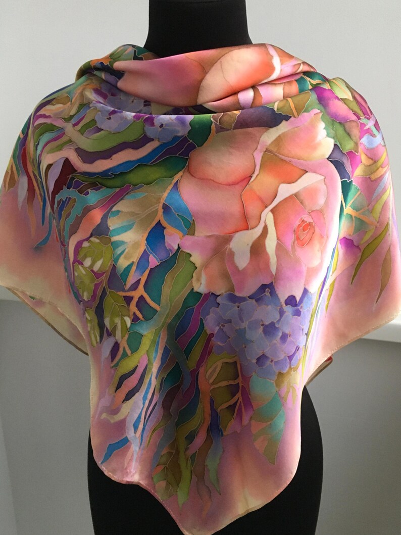 May include: A square silk scarf featuring a hand-painted floral design with large pink roses, clusters of purple hydrangeas, and green foliage. The fabric displays a vibrant color palette of pink, purple, green, and blue tones with defined outlines.