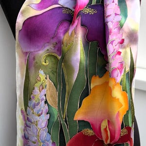 K&ouml;nnte beinhalten: Ein rechteckiger Seidenschal mit handgemaltem Blumenmuster, das leuchtend violette, orangefarbene und rosafarbene Schwertlilien, Lavendelst&auml;ngel und gr&uuml;nes Blattwerk zeigt. Unten links ist eine kleine braune Schnecke zu sehen. Der Schal hat einen helllila Rand.