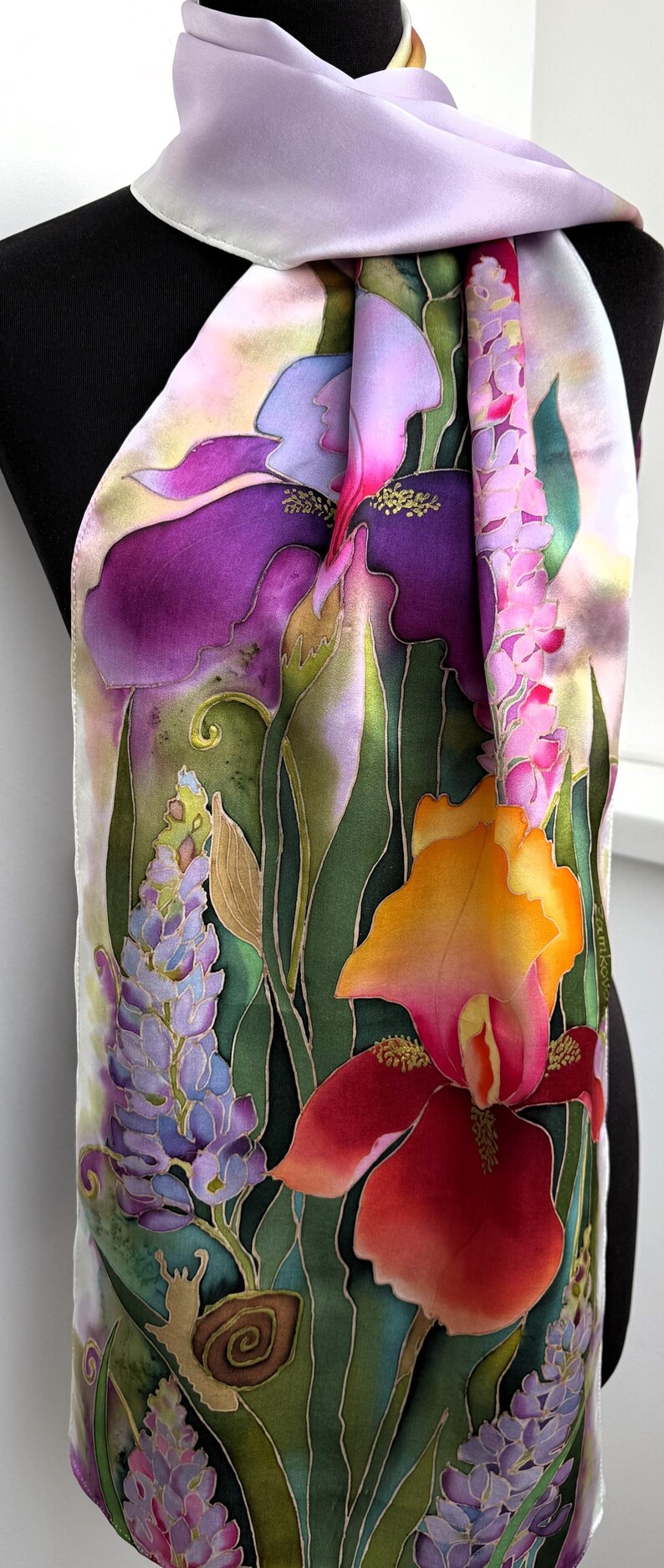 K&ouml;nnte beinhalten: Ein rechteckiger Seidenschal mit handgemaltem Blumenmuster, das leuchtend violette, orangefarbene und rosafarbene Schwertlilien, Lavendelst&auml;ngel und gr&uuml;nes Blattwerk zeigt. Unten links ist eine kleine braune Schnecke zu sehen. Der Schal hat einen helllila Rand.