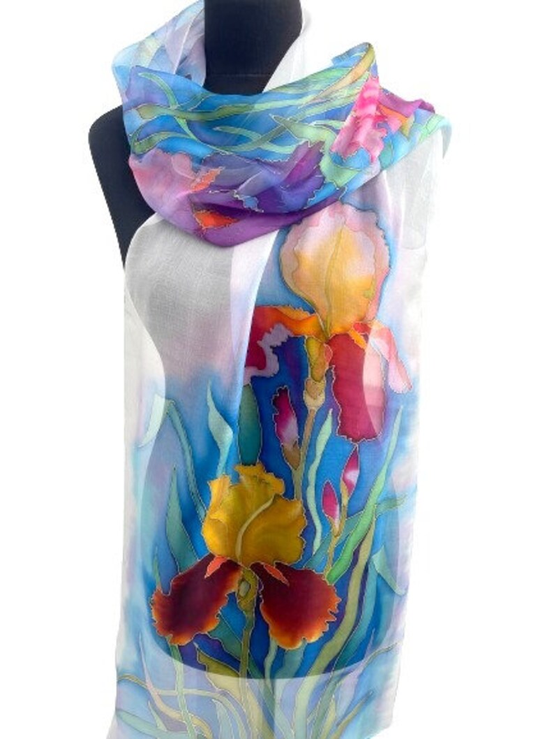 May include: A lightweight, semi-sheer scarf featuring a hand-painted floral design of vibrant irises in shades of yellow, deep red, and purple. The background consists of soft blue and white watercolor-style washes with green foliage accents.