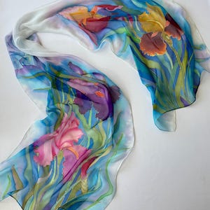 May include: A long, rectangular silk scarf featuring a hand-painted floral design of purple, pink, and yellow irises with green stems and leaves. The background is a blend of light blue and white, creating a soft, flowing, and artistic appearance.