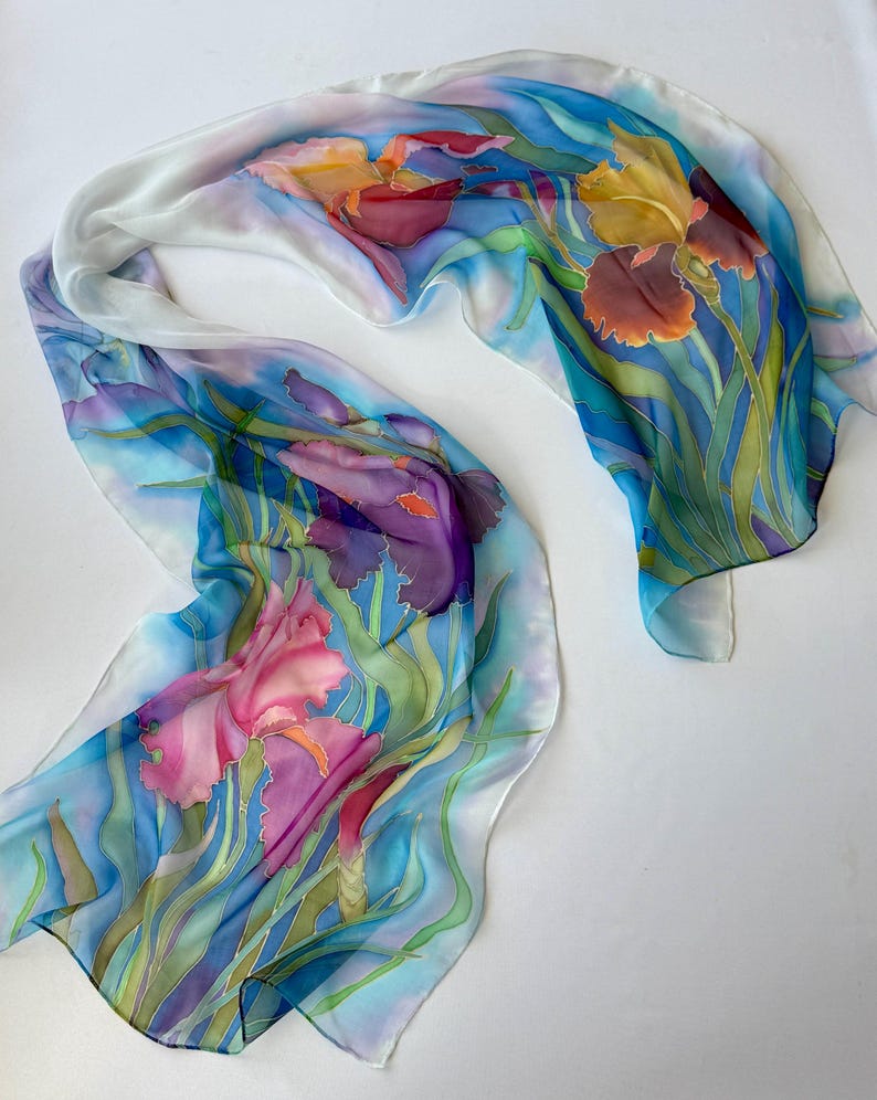 May include: A long, rectangular silk scarf featuring a hand-painted floral design of purple, pink, and yellow irises with green stems and leaves. The background is a blend of light blue and white, creating a soft, flowing, and artistic appearance.