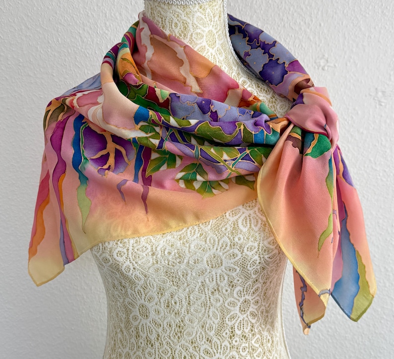 May include: A square silk scarf featuring a hand-painted design with a vibrant color palette of pink, purple, blue, and green. The pattern includes abstract flowing shapes and organic leaf-like motifs with defined outlines, draped around a mannequin.