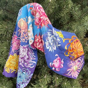 May include: A silk scarf with a blue background and a colorful floral pattern. The flowers are painted in shades of pink, purple, yellow, and orange. The scarf is draped over a pine tree branch.