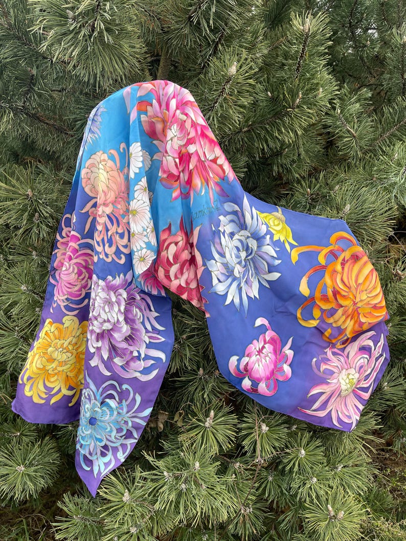 May include: A silk scarf with a blue background and a colorful floral pattern. The flowers are painted in shades of pink, purple, yellow, and orange. The scarf is draped over a pine tree branch.